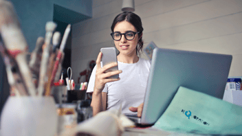 mulher de óculos feliz olhando para o celular e utilizando o computador